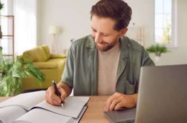 Gülümseyen erkek öğrenci dizüstü bilgisayarında oturuyor ve çevrimiçi ders sırasında not defterine not alıyor. Yetişkin öğrenci çevrimiçi çalışıyor ve oturma odasında oturan öğretmenleri uzaktan dinliyor..