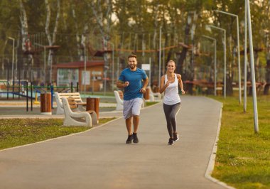 Parkta spor kıyafetleri içinde koşan mutlu, güler yüzlü bir çift spor antrenmanı yapıyor. Genç bir adam ve bir kadın dışarıda koşuyor. Doğada egzersiz, egzersiz ve sağlıklı yaşam tarzı konsepti..
