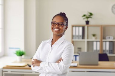 Ofiste poz veren başarılı, gururlu ve pozitif Afro-Amerikan iş kadını portresi. Beyaz gömlekli ve gözlüklü mutlu, esmer kadın kollarını kavuşturup kameraya gülümsüyor..