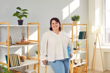 Oturma odasında elinde klasörle bekleyen ve gülümseyen şişman bir kadının portresi. Şişman, neşeli bir kız kameraya olumlu bakıyor. Vücut pozitif konsept.