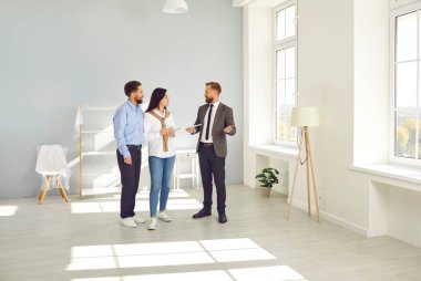 Elinde belgeler olan takım elbiseli yakışıklı sakallı bir emlakçı yeni bir ev almadan önce genç bir çifte tavsiye veriyor. Modern güneşli bir dairede üç kişi duruyor..