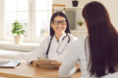 Steteskop takan gülümseyen doktorun portresi randevu ile rapor tutuyor ve danışmanlık yapıyor. Bir kadın klinikteki muayene sırasında ofisinde oturup doktorunu dinliyor.