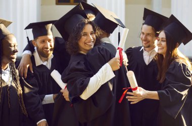 Mutlu gülümseyen çeşitli öğrenci grupları açık havada diplomaları ellerinde tutarak mezuniyetlerini kutluyorlar. Üniversite kampüsünde mezuniyet elbisesi içinde halka halinde duruyorlar..