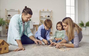 Genç bir aile evdeki oturma odasında iki çocuk ve bir kızla yerde oyun oynuyor. Gülümseyen ebeveynler çocuklarıyla yapboz topluyor. Aile ve insanlar kavramı.