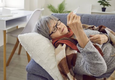 Yaşlı kadın üşütmüş. Grip hastası yaşlı bir kadın ateşini ölçüyor. Sıcak ekoseli ve atkılı yaşlı kadın oturma odasında kanepede uzanıyor ve termometreye bakıyor.