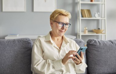 Evde akıllı telefon kullanarak boş zamanların tadını çıkaran üst düzey bir kadın. İnternet eğlencesine katılıyor, siber ortamda dinleniyor ve son yıllarında bağlantıda kalıyor..