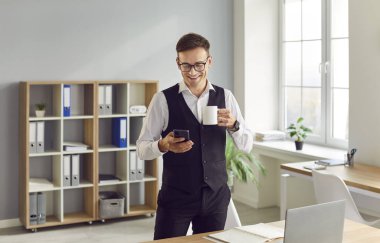 Genç, çekici, gülümseyen bir iş adamının portresi iş yerinin yanında elinde bir fincan çay ya da kahveyle duruyor ve cep telefonuna bakıyor ve mesajlarını okuyor ve gülümsüyor..