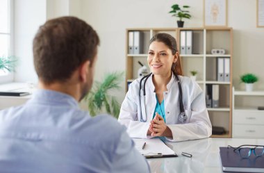 Dost canlısı bayan doktor masasında erkek hastasıyla oturuyor. Gülümseyen genç doktor ya da beyaz önlüklü pratisyen kadın hastaya tıbbi danışmanlık yapıyor. Doktor atama konsepti