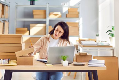 Depoda çalışan kendine güvenen bir bayan küçük işletme sahibi stokları ve envanterleri kontrol ediyor iş yerindeki masada oturuyor ve müşteriye depoda sipariş hazırlıyor. Teslimat ve depo işi konsepti.