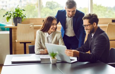Genç gülümseyen iş adamı iş yerinde otururken iş projesini bilgisayarında meslektaşlarına gösteriyor. Toplantıda gülümseyen insanlar şirket maliyesini tartışıyorlar..