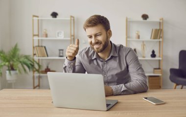 Mutlu genç iş adamı ya da şirket çalışanı evde oturuyor, dizüstü bilgisayara bakıyor, iş görüşmesi yapıyor ya da internet üzerinden görüşme yapıyor, onaylıyor ve gülümsüyor.