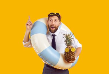 Ofis kıyafetleri giymiş, lastik yüzük takan heyecanlı komik bir adamın portresi ve stüdyo sarı arka planında izole edilmiş işaret parmağı olan taze bir ananas. Yaz tatili konsepti.