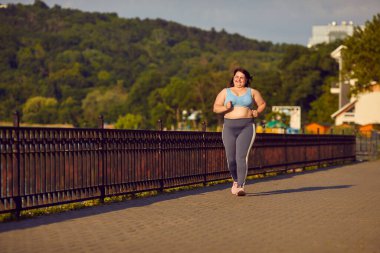 Şişman kadın hayatına egzersiz katıyor. Neşeli, güler yüzlü, şişman, şişman, spor sutyen ve spor pantolonlu kadın güneşli bir şehirde açık havada koşuyor ve köprüde koşuyor.