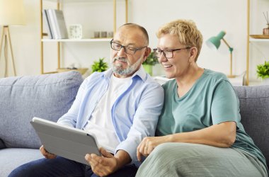 Rahat bir kanepede rahat rahat oturan yaşlı çift tablet kullanıyor, internette geziniyor, gülümsüyor ve modern teknolojiyle vakit geçiriyorlar. Yaşlılar evlerinde dijital anların tadını çıkarıyorlar.
