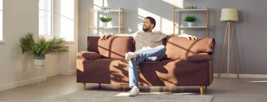 Genç adam evdeki rahat bir kanepede dinleniyor. Modern mobilyalarla, ev bitkileriyle ve büyük ışıklı pencerelerle dolu rahat bir oturma odasında, yumuşak kahverengi bir kanepede oturan mutlu bir adam. Sancak arkaplanı