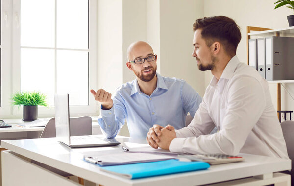 Two male business partners working with laptop together. Smiling young men in formal wear sitting at desk in modern office and discussing project. Leadership, partnership, collaborative teamwork