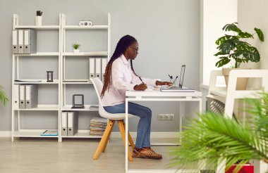 Kendine güveni tam, Afro-Amerikan bir kadının portresi. Günlük giysiler giymiş, çalışma masasında oturuyor ve modern ofiste ya da evinde bir dizüstü bilgisayarda çalışıyor..