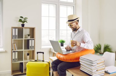 Ofisteki işyerinin masasında elinde plaj lastiğinde meyve suyu kokteyli ve dizüstü bilgisayarla yaz tatili için bilet ayırtan komik, mutlu bir iş adamı..