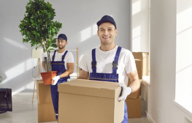 Portrait of smiling young man mover worker in uniform holding a cardboard box and looking cheerful at camera standing in empty new apartment. Move, relocation and moving service concept.