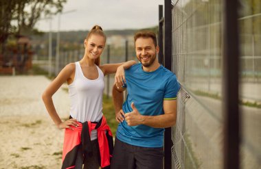 Spor kıyafetleri giymiş genç ve mutlu bir çift, şehir parkında kameralara bakıyor ve spor üzerine spor egzersizleri yapıyorlar. Doğa sporu, açık hava konsepti..