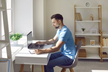 Genç adam evde dizüstü bilgisayar kullanırken notlar yazıyor. Ofiste masa başında çalışan, günlük kıyafetler giyen, odaklanmış bir adamın yan görünüm portresi. Evde ders çalışırken not defterine yazan erkek öğrenci