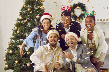 Neşeli genç, çok ırklı arkadaşların yeni yıl çanlarını yenmek için birlikte eğlenmelerinin portresi. Noel Baba şapkalı insanlar partideki konfeti altında poz veriyor, yüksek sesle ve neşeyle gülüyorlar..