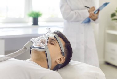 Adam hastane yatağında yatıyor, bir doktor ve hemşirenin gözetiminde oksijen maskesi takıyor. Astım tedavisi görüyor ve sağlık hizmetlerinin önemini vurguluyor..