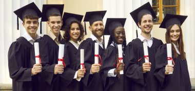 Gülümseyen mutlu, çok ırklı mezuniyet öğrencilerinin portresi üniversite mezuniyet elbisesi giymiş ve açık havada diploma tutuyorlar. Eğitim ve mezuniyet kavramı.