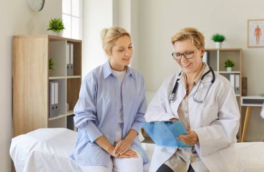 Beyaz önlüklü, olgun, deneyimli bir kadın doktor elinde bir dosya tutuyor ve modern klinikte muayene masasında oturan genç bir hastayla tıbbi sonuçları tartışıyor. Sağlık hizmetleri 