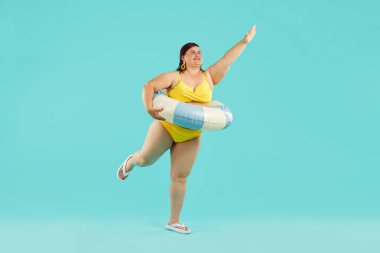 Sarı bikinili, kauçuk yüzüklü, yaz tatili gezisine çıkan ve mavi stüdyo arka planında eğlenen komik genç bir kadının vücut portresi. Tatil ve seyahat kavramı.