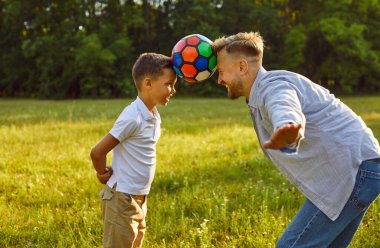 Mutlu baba ve çocuk yaz parkında eğleniyor. Neşeli baba ve oğul yeşil park çimlerinde oynuyorlar, alınlarıyla renkli futbol topunu tutuyorlar ve yere düşmesine izin vermiyorlar.