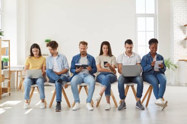 Sıraya dizilmiş dijital cihazlar kullanan çok kültürlü gençler. Çeşitli erkek ve kadınlar online olarak modern aletlerle eğleniyor, kitap okuyor ve sırada beklerken kağıda not alıyorlar.