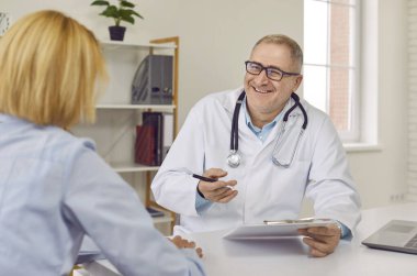 Dost canlısı gülümseyen olgun bir doktoru ziyaret eden hasta hastane ofisinde randevusuyla birlikte rapor tutuyor ve klinikte muayene sırasında bir kadına danışmanlık yapıyor..