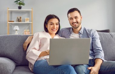 Mutlu gülümseyen genç çift evlerinde, oturma odasındaki rahat koltukta ellerinde dizüstü bilgisayarlarıyla birlikte monitör ekranına bakıyorlar. Mutluluk hissi, duygusal heyecan. Başarıyı kutla.