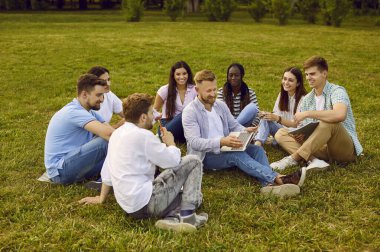 Çeşitli işletme öğrencileri ve arkadaşları doğa projesinde çalışıyor. Yaz parkında yeşil çimlerde oturan ve birlikte dizüstü bilgisayar kullanan bir grup mutlu genç insan.
