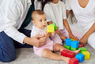 Küçük şirin bir bebeğin portresi yerde ailesiyle oturuyor ve oyuncak renkli eğitici küplerle. Genç mutlu ebeveynler evde çocukla piramit oynuyorlar. Aile ve insanlar kavramı.