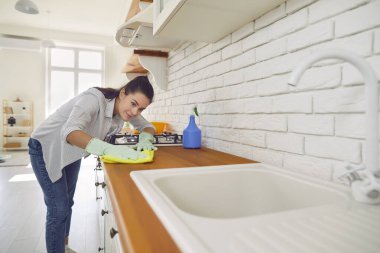 Kadın, evdeki mutfak mobilyalarını özenle temizleyip tozluyor. Eldivenlerini giyer, ev işlerine yoğunlaşır, evin temizliğini ve cazibesini arttırır..