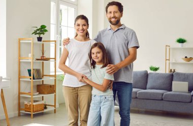 Gülümseyen mutlu, üç kişilik bir aile. Evlerinde kızlarıyla birlikte duruyorlar. Gülümseyen ebeveynler - anne, baba ve kızları oturma odasında sarılıyorlar ve kameraya neşeli görünüyorlar..