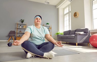 Aşırı kilolu komik ve kilolu genç bir kadın yoga minderinde Lotus pozunda kapalı gözlerle oturuyor, asma sana egzersizi yapıyor namaste jesti yapıyor, evde meditasyon yapıyor. Vücut pozitif konsept.