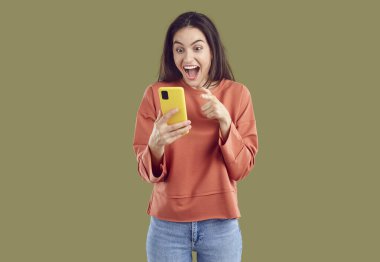 Kırmızı kazaklı ve kot pantolonlu mutlu, esmer kadının haki arka plandaki akıllı telefon ekranına bakmasına hayret ettim. O, ağzı açık ve geniş gözlerle gülüyor. İnanılmaz haber konsepti.