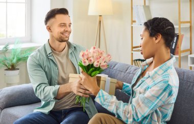 Evdeki kanepede oturan hediye kutusu ve çiçeklerle kadını tebrik eden bir erkek portresi. Mutlu genç çift bayramı kutluyor. Dikkat, tebrikler ve sevgililer günü konsepti.