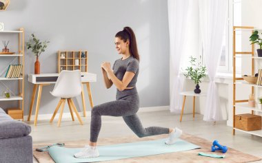 Aktif spor yapan genç bir kadın evde spor yapıyor. Oturma odasında yoga minderinde ön hamleler yapıyor. Spor giyim sektöründe atletik gülümseyen zayıf kız vücudunu güçlendiriyor, rutinini koruyor..