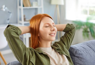 Genç, sakin, gülümseyen kızıl saçlı kadın rahat koltukta el ele tutuşuyor, mutlu kız işten sonra evde dinleniyor, tatilin tadını çıkarıyor, gözleri kapalı, stres yok.