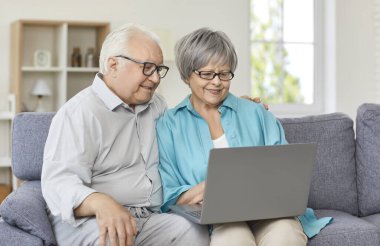 Birlikte dizüstü bilgisayar kullanan mutlu bir aile çifti. Kanepede oturup dizüstü bilgisayara bakarak gülümseyen yaşlı adam ve kadın. Emekli karı-koca videosu akrabalarını arıyor ya da internet şovu izliyor.