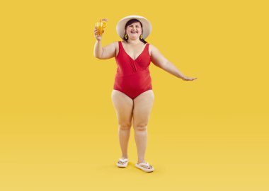 Kırmızı mayo ve plaj şapkalı, elinde portakal suyu kokteyli olan mutlu ve şişman bir kadının portresi. Sarı arka planda neşeli görünüyor. Yaz tatili ve tatil konsepti.