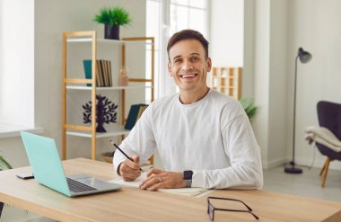 Ofiste dizüstü bilgisayarda çalışan genç çekici iş adamının portresi ya da evde planlanan günlük toplantı ve randevu kameraya neşeli görünüyor. Uzak ve serbest çalışma konsepti.