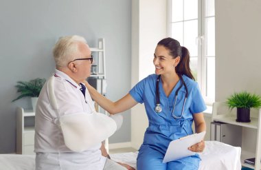 Kırık kollu gözlüklü, yaşlı, gri saçlı, gülen adam, ilgili hemşireyle konuşuyor, tıbbi kanepede oturuyor. Genç, güzel, destekleyici kadın doktor sağlıksız yaşlı adamı konsültasyonda rahatlatıyor.