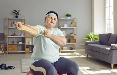 Spor egzersizleri yapan şişman, komik bir kadının portresi. Elinde lastik bantla evde oturmuş spor giyerek spor yapıyor. Spor, spor, ev eğitimi ve vücut pozitifliği.