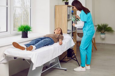 Dost canlısı, önlüklü, güler yüzlü kadın hemşire klinikte yatan küçük çocuk hasta çocuğa damardan damardan ilaç veriyor. İlaç, tedavi, vitamin terapisi konsepti