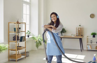 Evi elektrikli süpürgeyle, kulaklıkla temizleyen kadın ev işlerine neşeli bir ritim getiriyor. Hizmetçi portresi bulaşıcı pozitifliği yansıtıyor. Oturma odasının zeminini zarafetle süpürüyor..